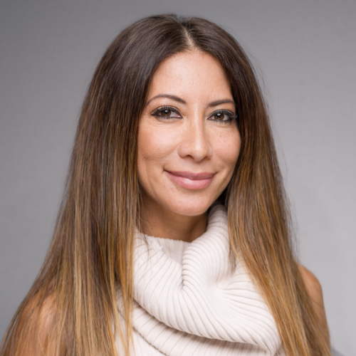 A portrait of a person with long, dark, wavy hair wearing a cream-colored turtleneck sweater. The person is smiling gently at the camera against a white background.