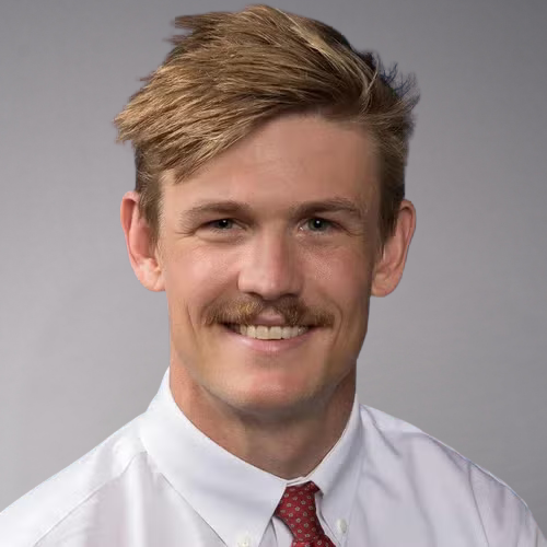 A man with short, styled blond hair and a mustache smiles while wearing a white dress shirt and a red tie with white polka dots.