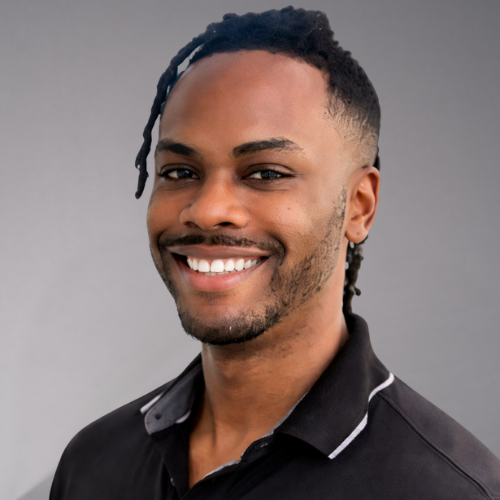 A man with braids and a black polo shirt smiles broadly, showing white teeth. The background is white.