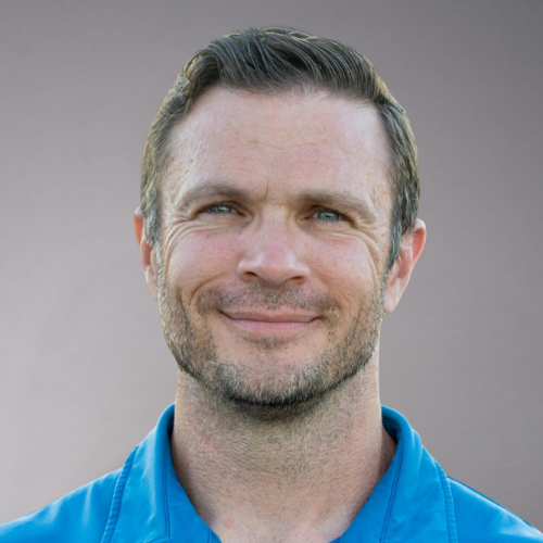 Close-up portrait of a man with short brown hair, blue eyes, and a beard, wearing a blue collared shirt.