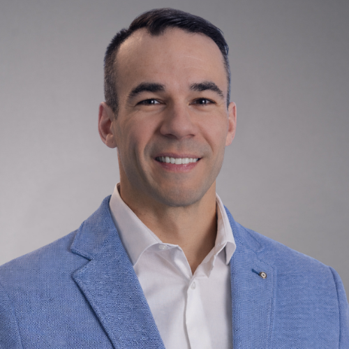 A man wearing a blue textured blazer over a white collared shirt smiles at the camera against a white background.